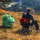 A man sits on a hill overlooking a forest with a smiling Bulbasaur nearby.